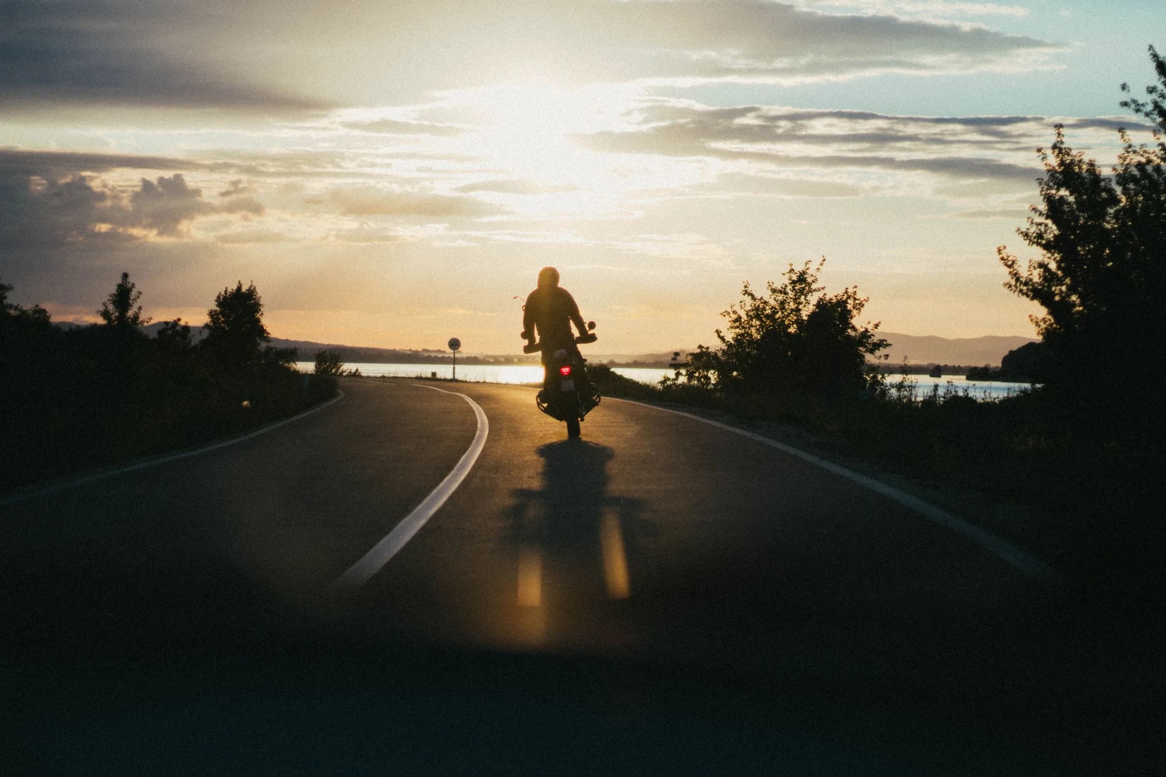 Motociclista de espaldas conduciendo hacia un atardecer en una carretera abierta